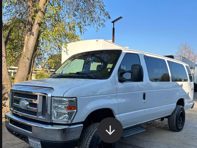 A white Ford E-350 4x4 passenger van with lifted suspension and all-terrain tires, parked in a gravel yard.