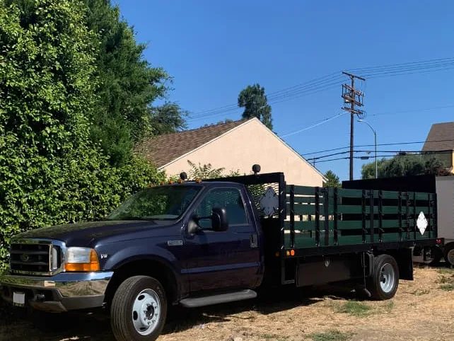 A dark blue Ford F-450 stakebed truck with dark green wood stake sides, parked on a dirt lot.