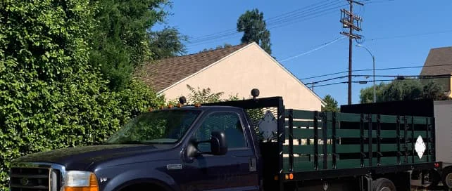 A dark blue Ford F-450 stakebed truck with dark green wood stake sides, parked beside a hedge and a house in daylight.