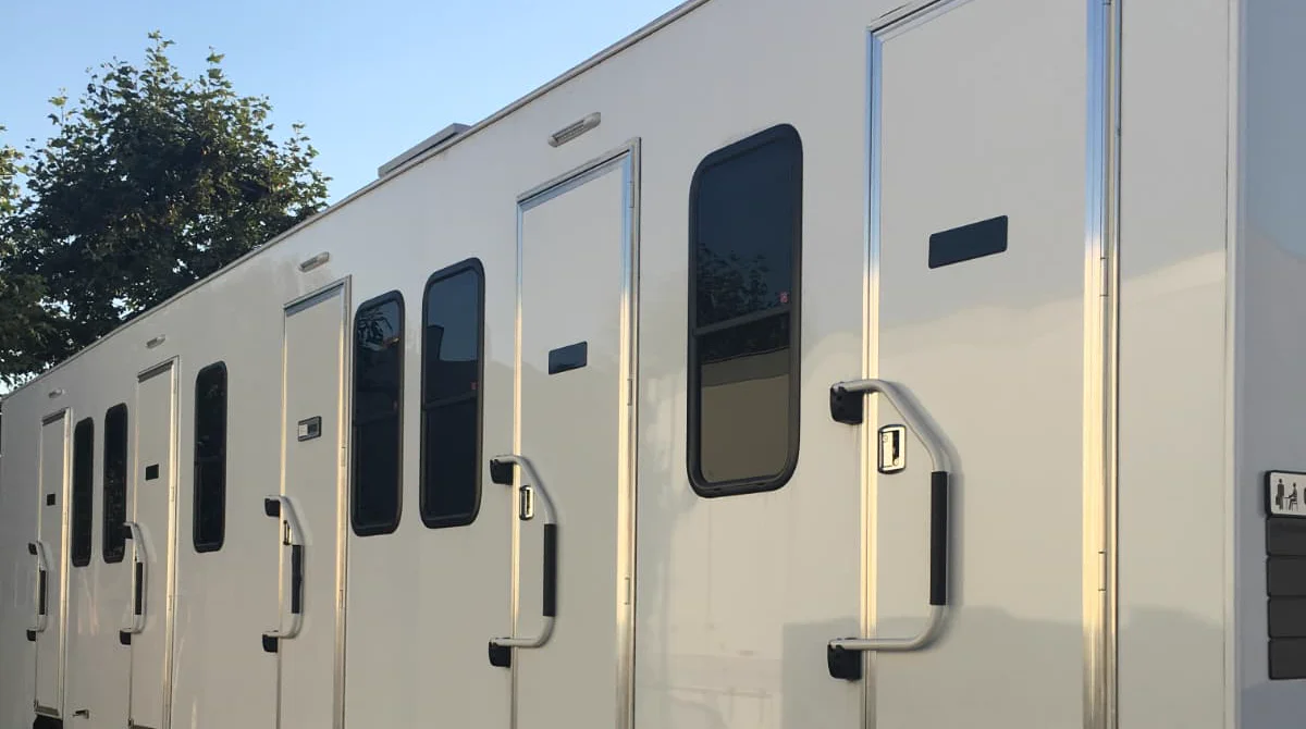 A row of doors and tinted windows along a white multi-room honeywagon trailer, lit by golden-hour sunlight, with a tree and blue sky above.