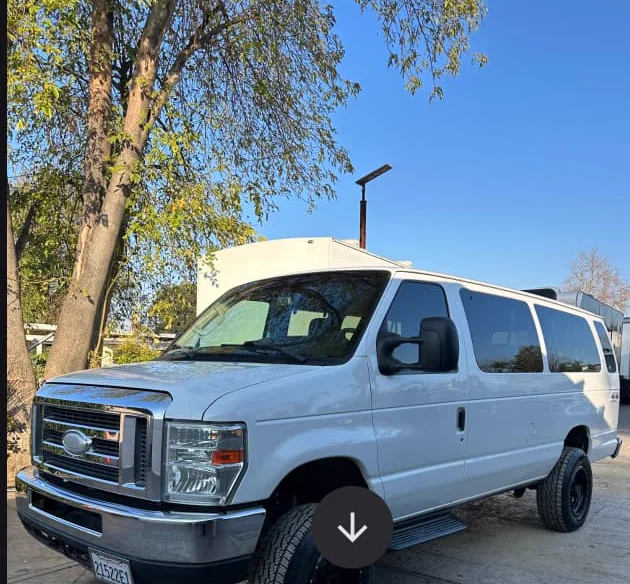 A white Ford E-350 4x4 passenger van with lifted suspension and all-terrain tires, parked under a tree in a gravel yard.