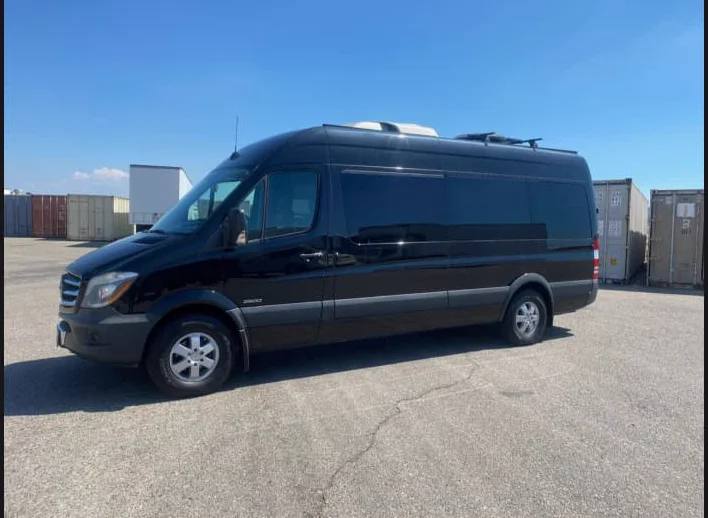 A black Mercedes-Benz Sprinter 2500 extended high-roof van, viewed from the front three-quarter angle, parked in a shipping-container yard under a clear blue sky.