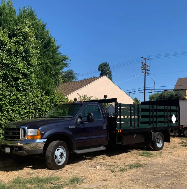 A dark blue Ford F-450 stakebed truck with dark green wood stake sides, parked on a dirt lot in daylight, with a large hedge and a house in the background.